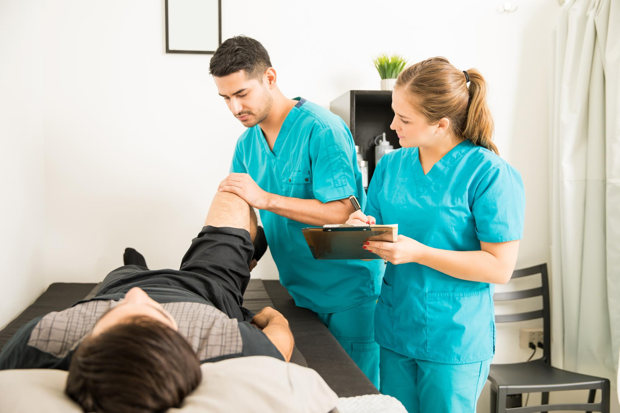 young-physiotherapist-examining-customer-s-leg-while-colleague-noting-clipboard-hospital